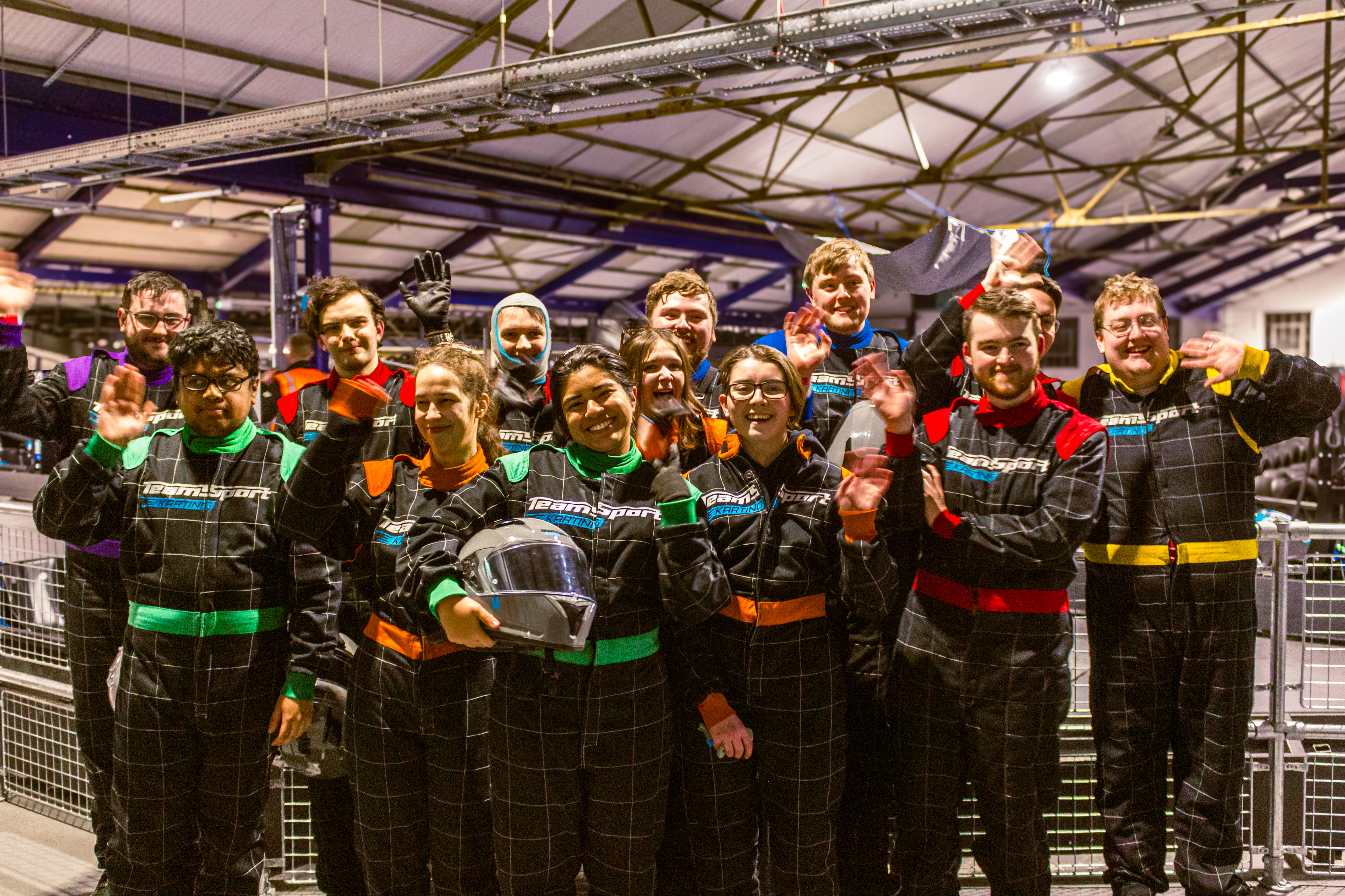 A group of students at a go kart course wearing overalls, holding helmets and making peace signs