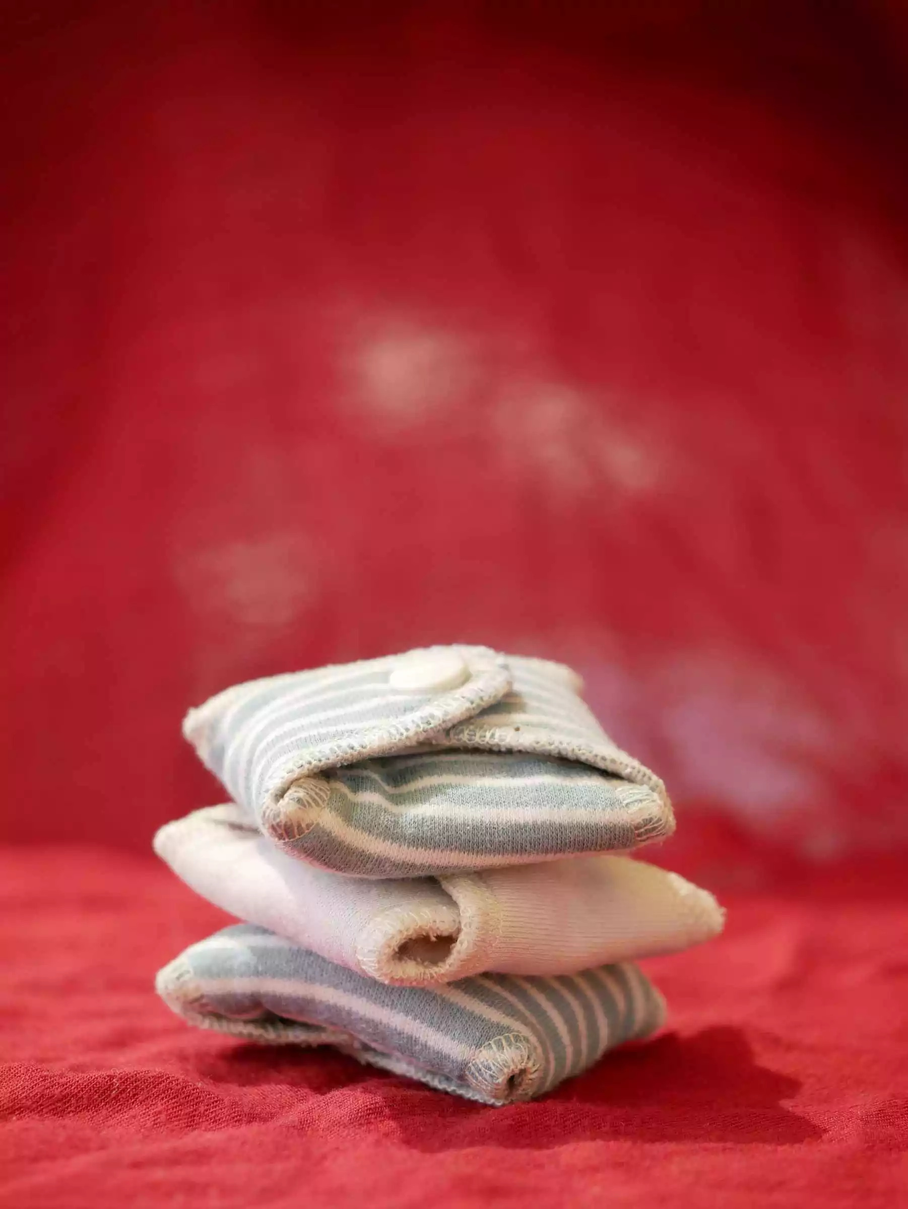 Photograph of three floded reusable sanitary pads in front of a red backdrop. Photo by Vulvani – www.vulvani.com’
