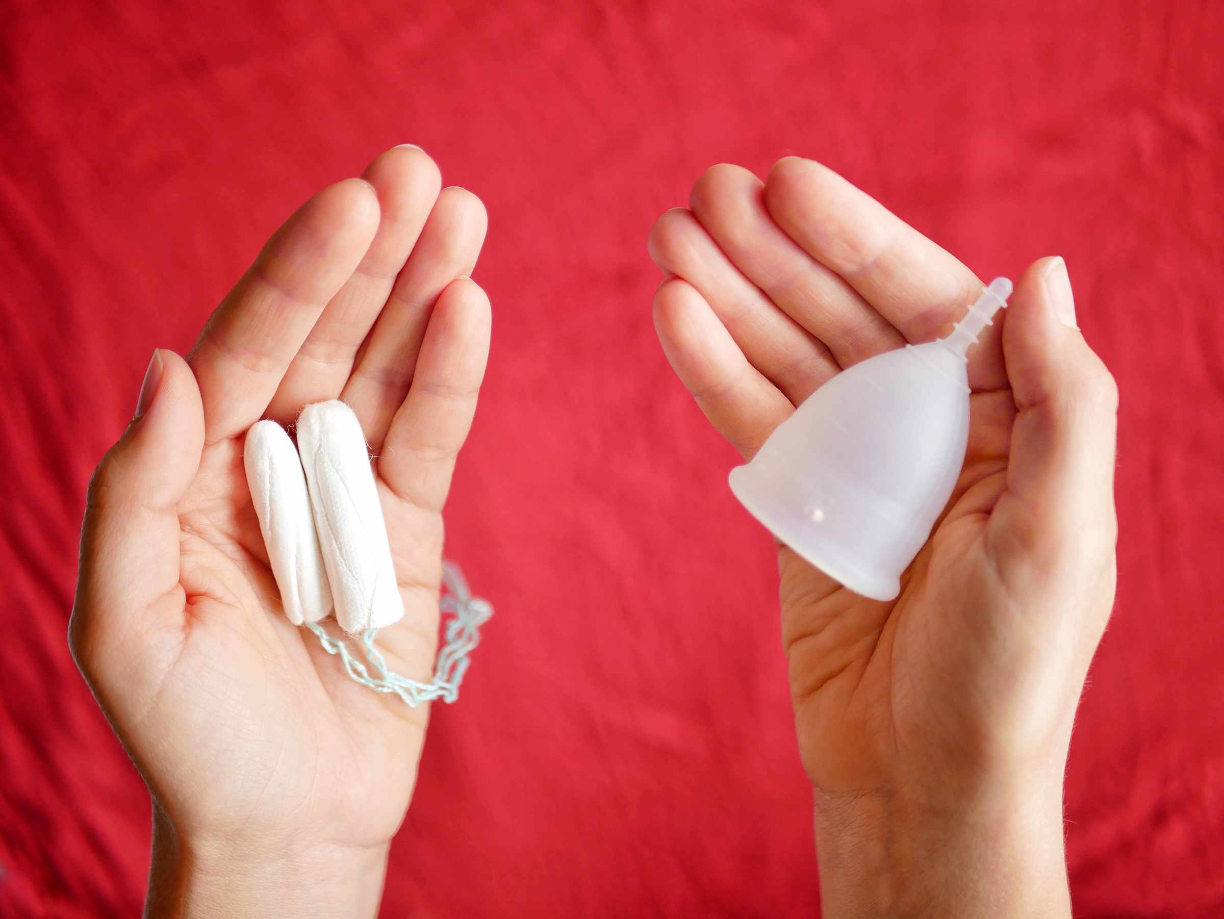 Photo of two hands palm up with a red backdrop, one with two tampons in, the other a menstrual cup in. Photo by Vulvani - www.vulvani.com