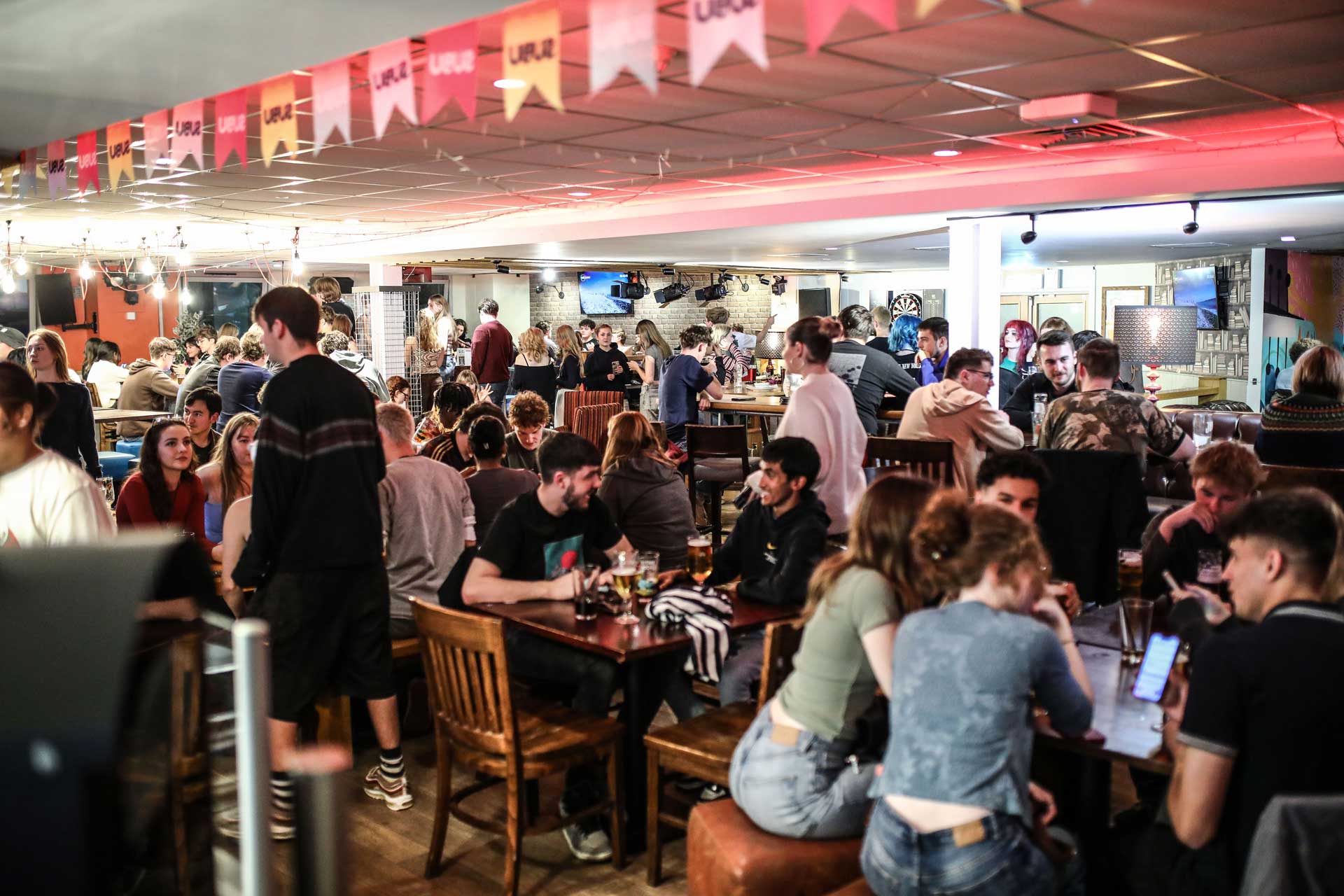Students sitting in Dylan's Bar and Kitchen, it's busy and looks cosy with warm red lighting and wooden tables and chairs